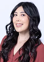 Smiling person with long dark wavy hair, wearing a burgundy blouse against a light gray background.
