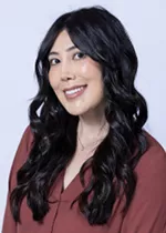 Smiling person with long dark wavy hair, wearing a burgundy blouse against a light gray background.