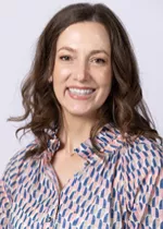Smiling person with shoulder-length wavy brown hair, wearing a patterned blouse against a light gray background.