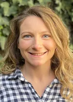 Portrait of a smiling woman with wavy blonde hair wearing a blue-and-white checkered shirt, with greenery in the background.