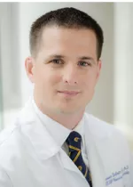 Head-and-shoulders portrait of a man wearing a white medical coat, white shirt, and patterned tie, facing the camera against a softly blurred light background.