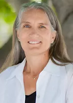 Headshot of a smiling woman with long gray hair, wearing a white collared shirt over a black top, photographed outdoors against a softly blurred green background.