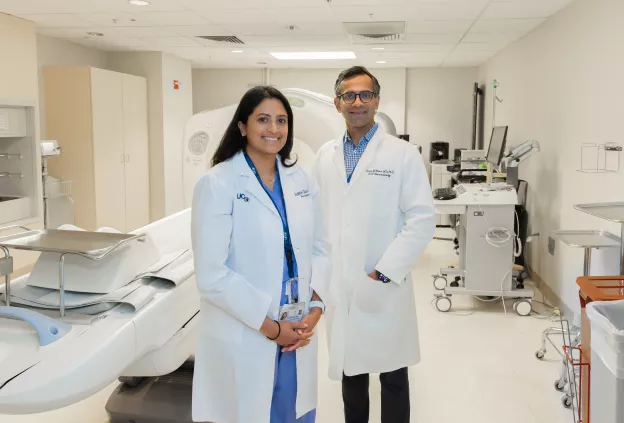 Two physicians in white lab coats standing in a hospital imaging room with a CT scanner and medical equipment in the background.