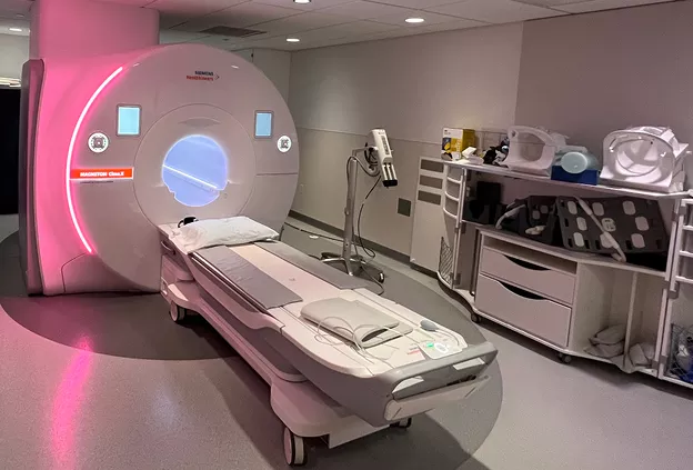 MRI scanner in a hospital imaging room with a patient table extending from the circular scanner, surrounded by medical equipment and storage carts.