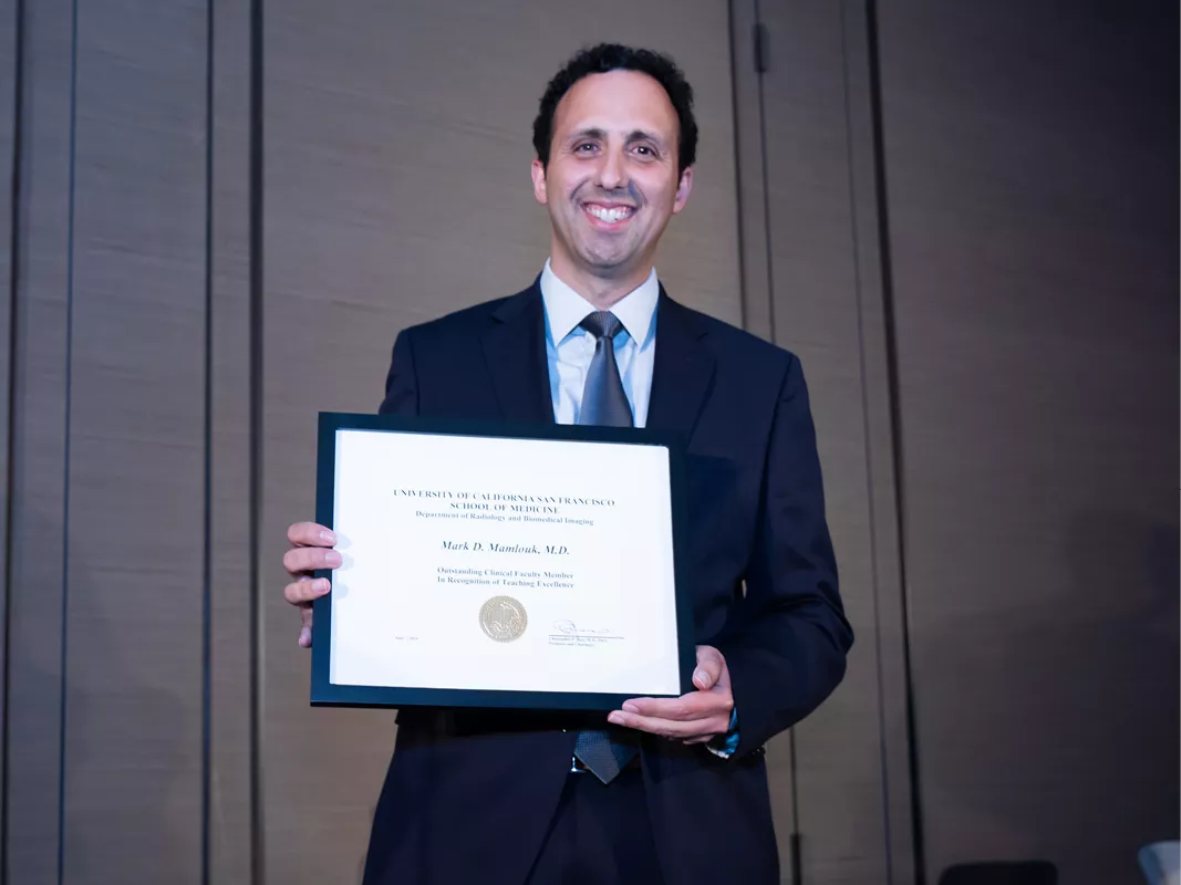 Person in a suit standing indoors and smiling while holding a framed certificate toward the camera.