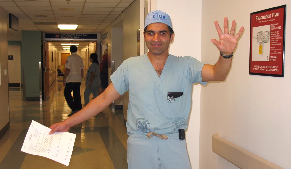 Rajiv Sawhney wearing light blue scrubs and a surgical cap stands in a hospital hallway, holding papers in one hand and waving with the other. Several people are visible in the background walking down the corridor.