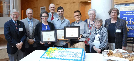 Group of eleven people posing indoors behind a table with a decorated cake that reads Celebrating 10 Years Surbeck Scholars. Three individuals in the front hold framed certificates while others stand around them smiling."
