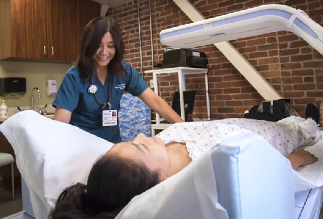 technologist assisting a patient lying on a padded table during a body composition x-ray scan, with the scanning arm positioned overhead in a clinical room with brick walls
