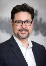Portrait of Christopher Hess wearing glasses and a dark blazer, smiling slightly against a blurred gray background.