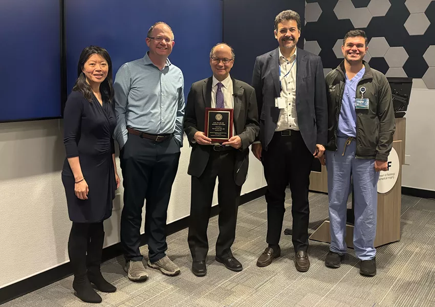 Group of five people standing together in a conference room. The person in the center is holding an award plaque while the others stand beside them, smiling. A podium and presentation screens are visible in the background, suggesting a formal recognition or lecture event.