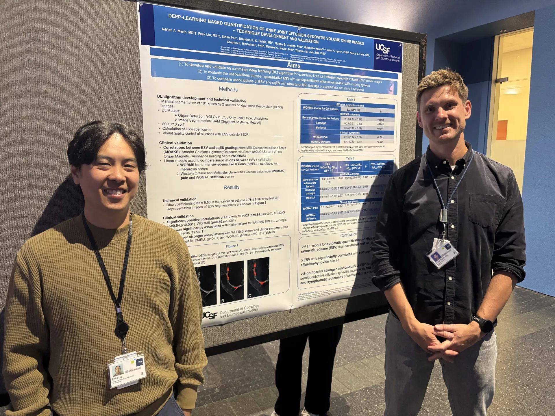 Two researchers stand on either side of a large scientific poster titled “Deep-Learning Based Quantification of Knee Joint Effusion–Synovitis Volume on MR Images: Technique Development and Validation” displayed on a poster board at a conference. Both are wearing lanyards with identification badges. The researcher on the left wears a tan sweater and smiles toward the camera, while the researcher on the right wears a dark button-up shirt and stands with hands clasped. The poster includes sections such as Aims
