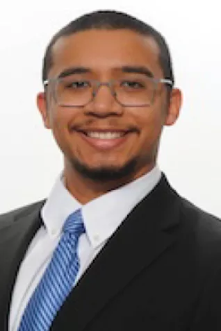 Professional headshot of a smiling person wearing glasses, a dark suit, white shirt, and light blue tie against a white background.