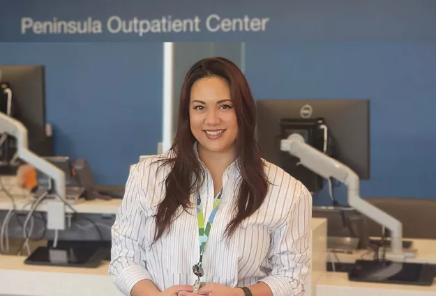 Amy Acuna stands smiling in the Peninsula Outpatient Center reception area. She is wearing a white striped blouse and staff badge, with registration desks and computer workstations visible in the background. The image highlights her role in supporting patient care and operations at the outpatient center.