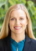 Professional headshot of a smiling woman with long blonde hair, facing the camera. She is wearing a dark blazer over a teal blouse. The background is softly blurred with green foliage, creating a warm and approachable appearance while maintaining a polished, professional look suitable for a faculty or staff profile.