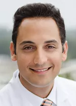 Professional headshot of a smiling man with dark hair, wearing a white dress shirt and a patterned tie. He is facing the camera against a softly blurred outdoor background. The portrait has a clean, approachable appearance suitable for a faculty, physician, or professional profile.