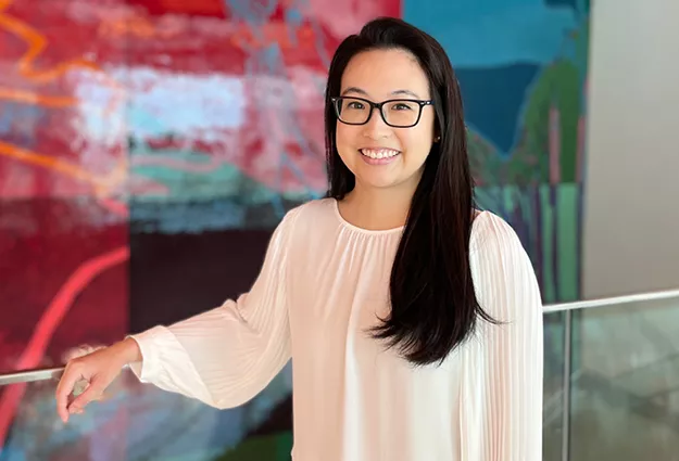 A woman with long dark hair and glasses smiles while standing indoors beside a railing. She wears a light-colored blouse, with a colorful abstract mural softly blurred in the background.