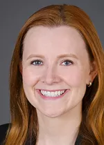 Professional headshot of a smiling woman with long red hair, facing the camera. She is wearing a dark blazer and small stud earrings. The portrait is cropped closely around her face and shoulders and set against a neutral gray background, creating a polished and professional appearance suitable for a faculty or staff profile.