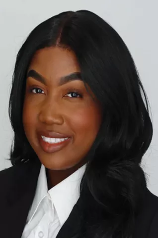 Professional headshot of a smiling person with long black hair wearing a dark blazer and white collared shirt against a white background.