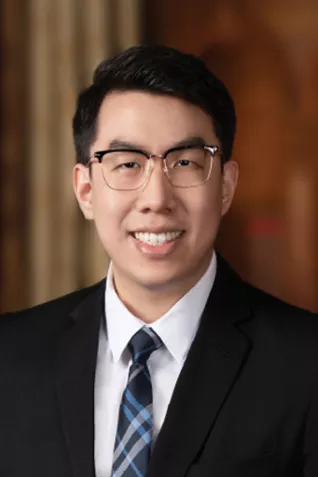 Professional headshot of a smiling person wearing glasses, a dark suit, white shirt, and blue plaid tie against a warm blurred indoor background.