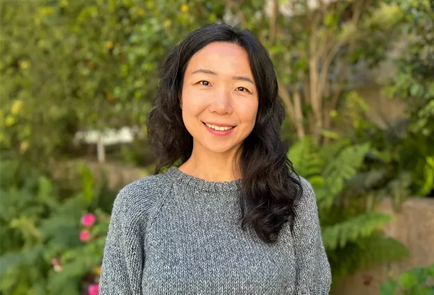 Portrait of a woman with long dark hair wearing a gray knit sweater, standing outdoors and smiling at the camera. Trees, plants, and soft natural light create a blurred garden background.