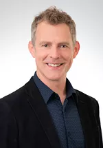 Professional headshot of a smiling man with short, curly light-brown hair wearing a dark blazer over a blue collared shirt. He is facing the camera against a plain light gray background.