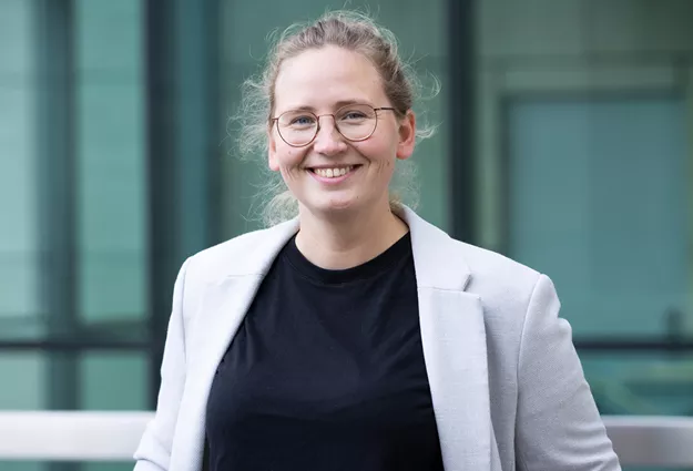Portrait of Katharine Ziegler wearing glasses, a black top, and a light-colored blazer, smiling at the camera. She stands outdoors in front of a modern building with a softly blurred background.