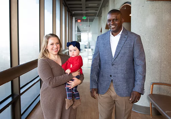 UCSF radiologist Kevin McGill stands with patient Maryana Kessel and her young child in a hospital hallway. Kessel holds her child while McGill stands beside them, smiling at the camera. The image highlights the partnership between physician and patient in delivering compassionate, family-centered care.