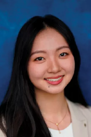 Professional headshot of a smiling person with long black hair wearing a light blazer and white top against a blue studio background.
