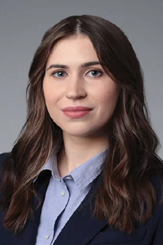 Professional headshot of a person with long wavy brown hair wearing a navy blazer and light blue button-down shirt against a gray-blue background.