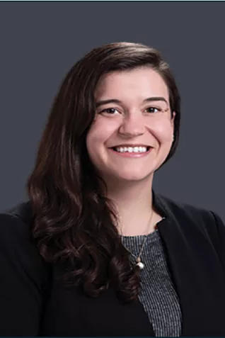 Professional headshot of a smiling person with long dark wavy hair wearing a black blazer, gray top, and necklace against a dark gray background.