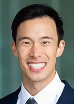 Professional headshot of a smiling man with short dark hair, wearing a dark suit jacket, white dress shirt, and tie. He is facing the camera against a softly blurred green background. The portrait is well lit and polished, creating a professional appearance suitable for a faculty, physician, or leadership profile.