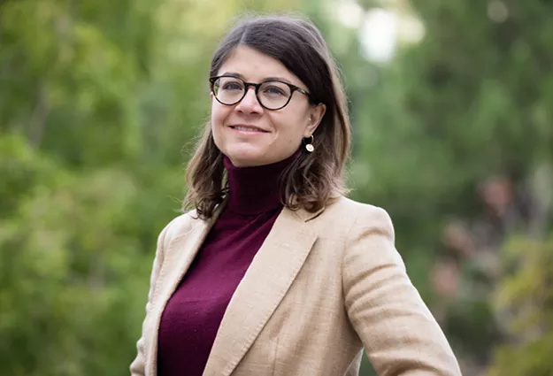 Portrait of Virginie Kreutzinger wearing glasses, a burgundy turtleneck, and a tan blazer, standing outdoors and looking toward the camera. Trees and greenery form a softly blurred background.