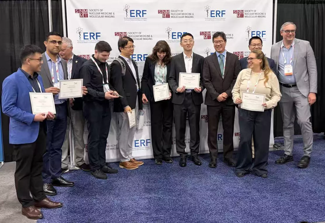 Group of award recipients and presenters posing for a photo at a professional conference. Several individuals stand in front of an SNMMI and ERF branded backdrop, holding certificates and wearing conference badges. The group is gathered on a blue carpeted stage area inside a convention center, with promotional banners displayed on either side. Most participants are smiling or looking toward the camera during the recognition ceremony.