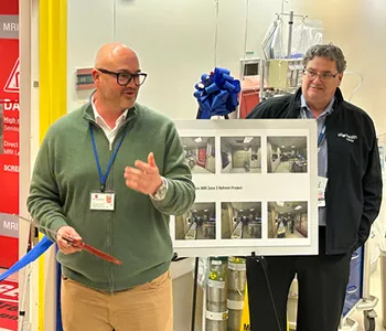 A staff member speaks during a hospital ribbon-cutting ceremony while standing beside another colleague and a display board showing project photos. A blue ribbon and bow decorate the entrance behind them.
