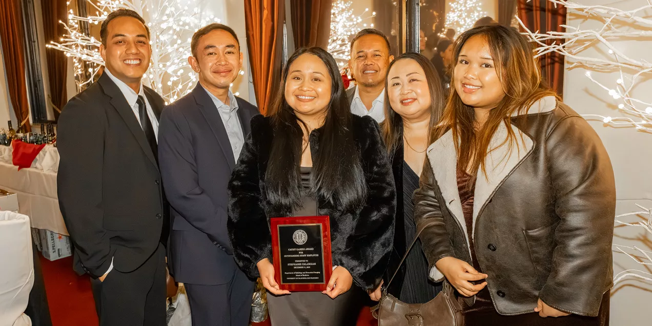 Group photo of six people dressed in formal attire at an indoor awards event. The person in the center holds a recognition plaque while the group smiles for the camera in front of decorative white lighted branches.