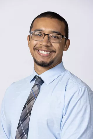 Smiling person wearing glasses, a light blue dress shirt, and a gray plaid tie against a light gray background.