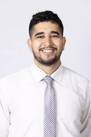 Smiling person with short curly dark hair and trimmed facial hair, wearing a white dress shirt and a striped gray tie against a light gray background.