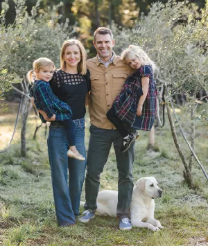 Family of four posing outdoors among olive trees with their dog, with the parents holding their two young daughters.