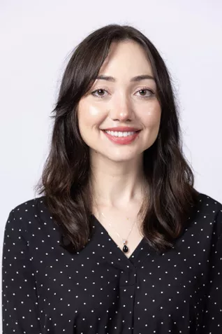Smiling person with shoulder-length dark brown hair, wearing a black blouse with a subtle dot pattern against a light gray background.