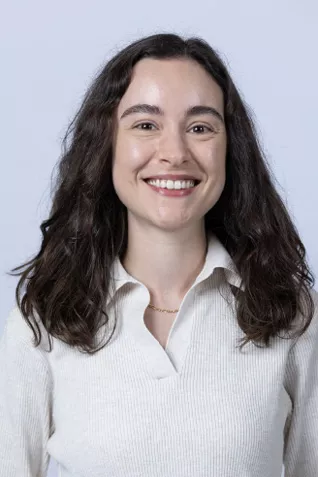 Smiling person with long dark wavy hair, wearing a white collared blouse against a light gray background.