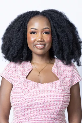 Smiling person with natural curly hair, wearing a pink patterned top and a necklace against a light gray background.