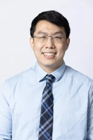 Smiling person wearing glasses, a light blue dress shirt, and a blue plaid tie against a light gray background.