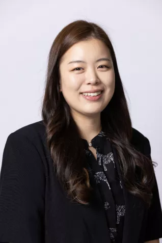 Smiling person with long dark hair, wearing a black blazer over a patterned blouse against a light gray background.