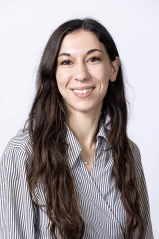 Smiling person with long dark hair, wearing a striped button-up shirt against a light gray background.