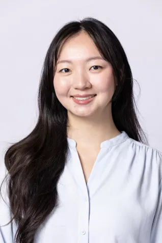 Smiling person with long dark hair, wearing a light blue blouse against a light gray background.
