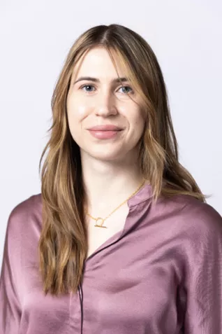 Person with long blonde hair, wearing a mauve satin blouse and a necklace against a light gray background.