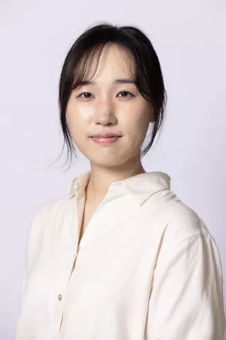 Person with dark hair pulled back, wearing a cream-colored blouse against a light gray background.