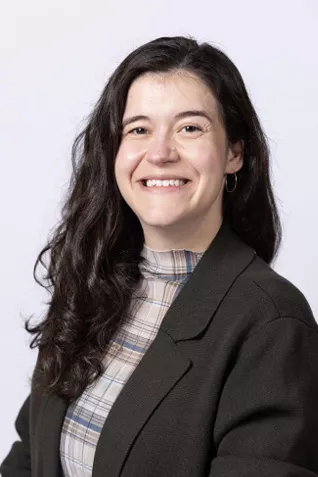 Smiling person with long dark hair, wearing a dark blazer over a plaid mock-neck top against a light gray background.