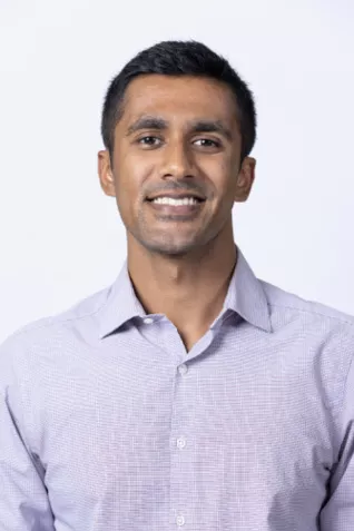 Smiling person with short dark hair, wearing a light lavender button-up shirt against a light gray background.
