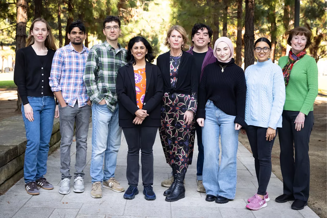 Group photo of nine lab members standing together on a tree-lined walkway outdoors.